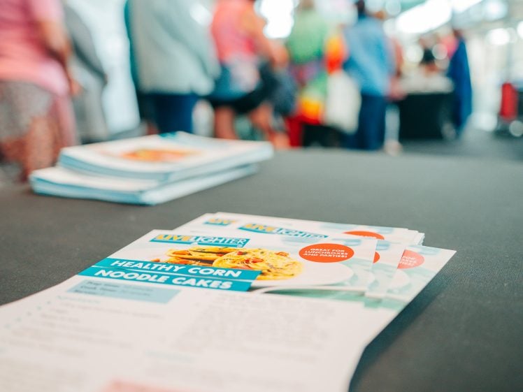 Image of A close-up of a stack of leaflets featuring a recipe for healthy corn noodle cakes on a table, with people standing in a queue blurred in the background.