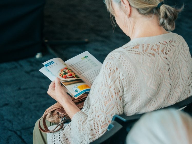Image of An older woman with grey hair sits and reads a magazine or recipe booklet, which features a photo of a salad. She wears a light, lacy, cream-coloured top and holds a brown handbag on her lap. The background is out of focus.