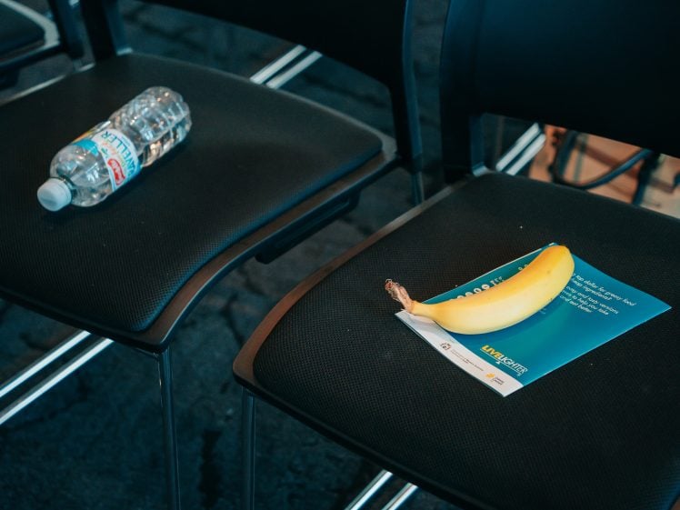Image of A plastic water bottle rests on one black chair, while a banana sits on a leaflet atop a neighbouring black chair. Both chairs are empty and part of a row.