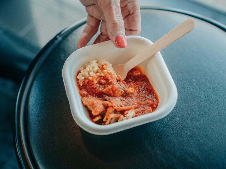 Image of A hand with red nail varnish holds a small bowl of rice and tomato-based stew with a wooden spoon, placed on a black circular surface.
