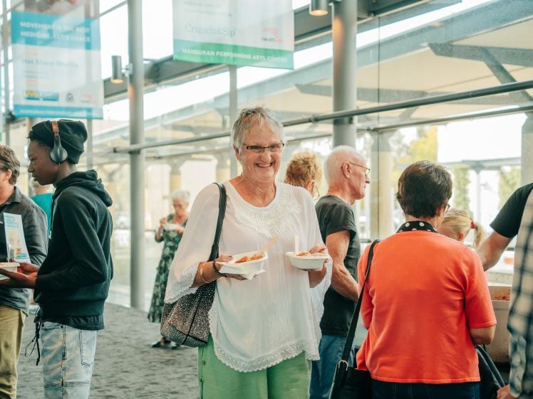 Image of A group of people socialise indoors at an event. A smiling older woman in a white blouse holds two plates of food, surrounded by others who are chatting and eating. The setting appears modern and well-lit with large windows.