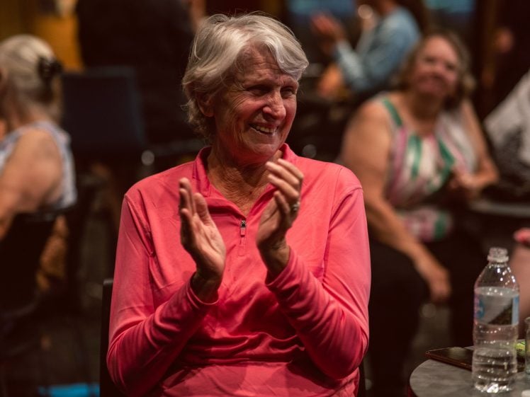Image of An older woman with grey hair, wearing a pink top, sits at a table and smiles while clapping. Other people are blurred in the background, and a water bottle is visible on the table in front of her.