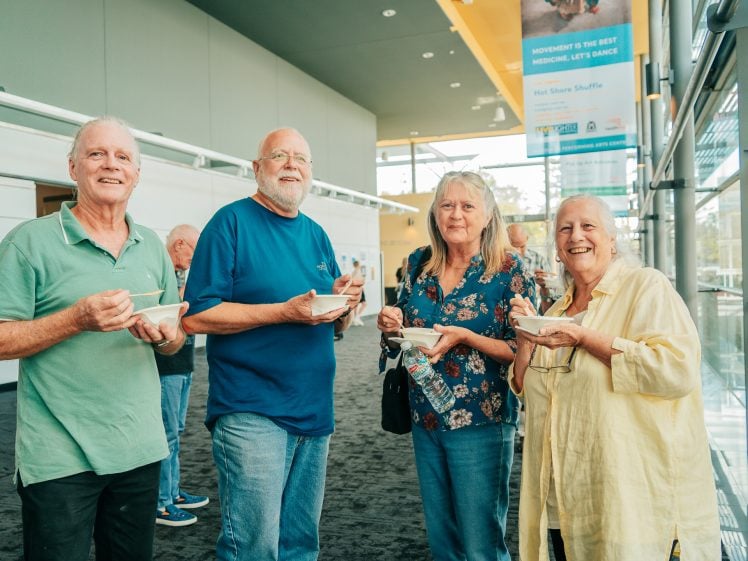 Image of Four older adults, two men and two women, stand smiling indoors while holding bowls of food. They appear to be at a social event in a bright, modern building with large windows.