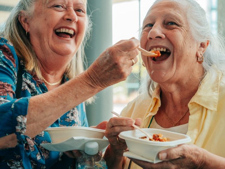 Image of Two older women smiling and laughing whilst eating food from bowls. One woman feeds the other with a fork. Both appear cheerful and are standing close together, enjoying their meal indoors.