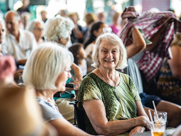 Image of A group of elderly people sit together at a lively indoor event. Two women in the foreground are smiling and talking, with drinks on the table. The background is filled with other attendees.