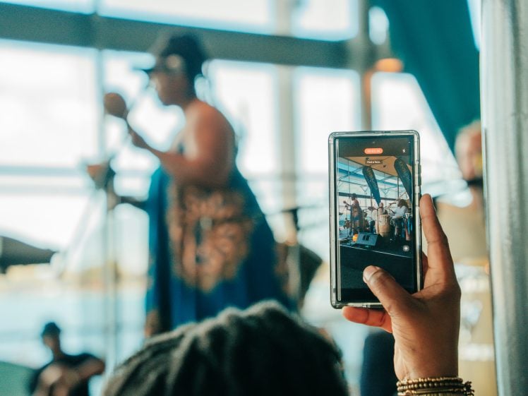 Image of A person holds up a mobile phone to record or take a photo of a singer performing on stage, with the singer blurred in the background and the phone screen showing a clear image of the performance.