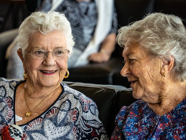 Image of Two elderly women with white hair sit closely together on a sofa, smiling and engaged in conversation. Both wear patterned blouses and gold earrings, creating a warm and friendly atmosphere.