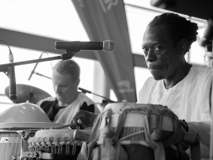 Image of Two musicians perform on stage. The foreground musician, in a white shirt, plays hand drums while looking towards the camera. Another musician in the background plays drums. Microphones are set up in front of them. The image is black and white.