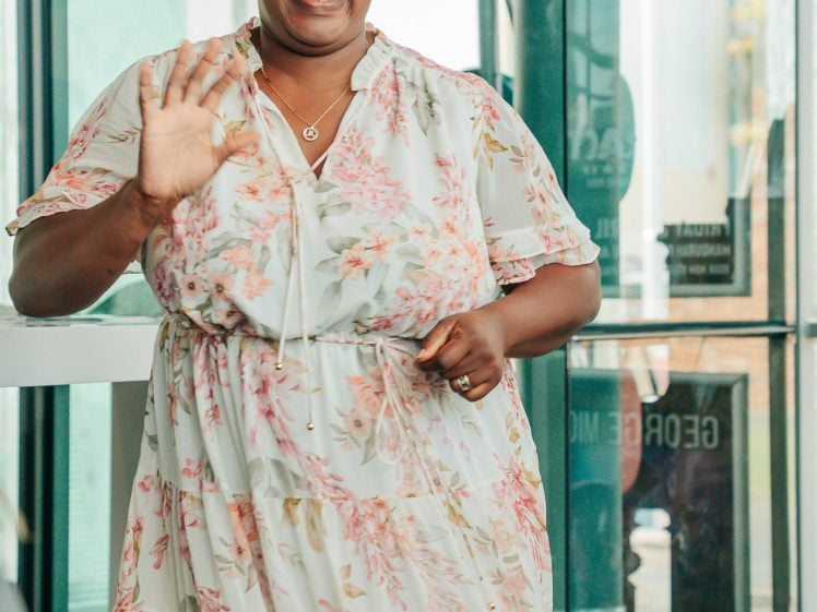 Image of A woman in a floral dress smiles and waves whilst standing indoors near large glass windows.