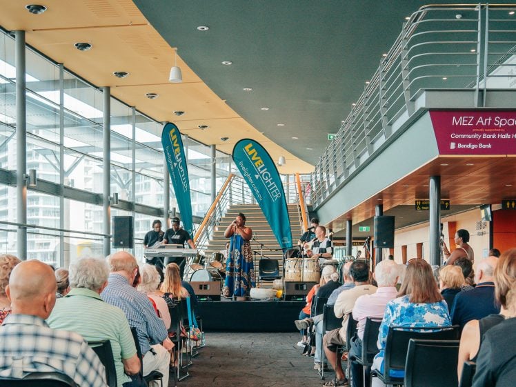 Image of A crowd watches a live indoor music performance at a modern venue with large windows, upstairs walkway, and MEZ Art Space sign. Two blue banners are on stage behind the musicians.