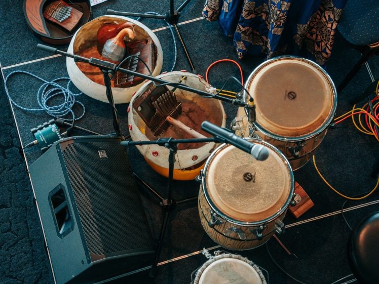 Image of Overhead view of various drums, gourd instruments, microphones, and audio cables on a stage floor next to a large black speaker and a chair draped with patterned fabric.