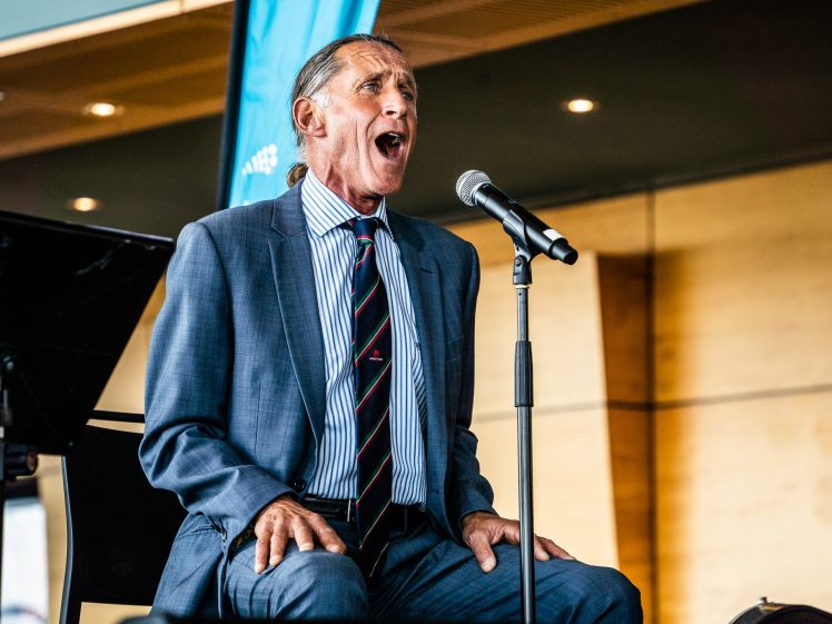 Image of An older man in a blue suit and striped tie sits on a chair, passionately singing or speaking into a microphone on stage in an indoor setting.