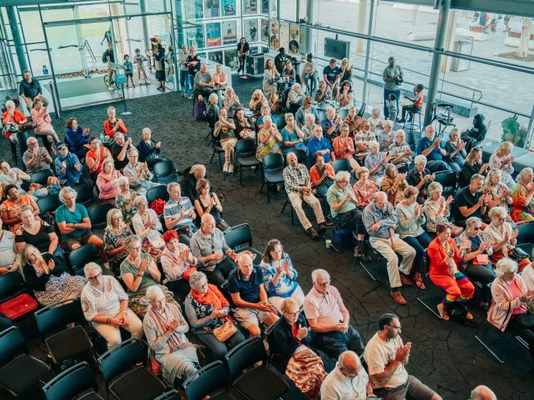 Image of A large group of people, mostly older adults, sit in rows of chairs inside a bright, modern building, facing the front. Some people are applauding, while others are talking or watching attentively.