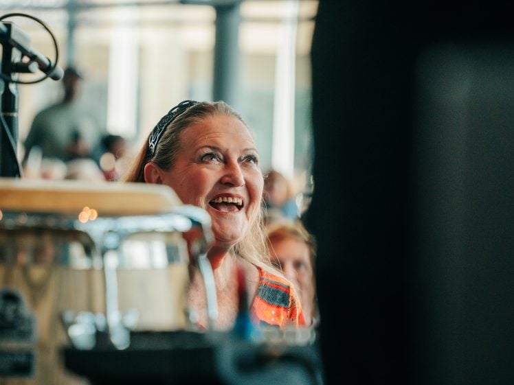 Image of A woman with long blonde hair smiles brightly whilst sitting indoors near a drum. The background is softly blurred, suggesting a lively event or performance.