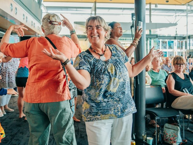 Image of A smiling woman in a blue patterned shirt and white trousers dances in a bright, indoor space filled with other people, some seated and some standing, enjoying the lively atmosphere.