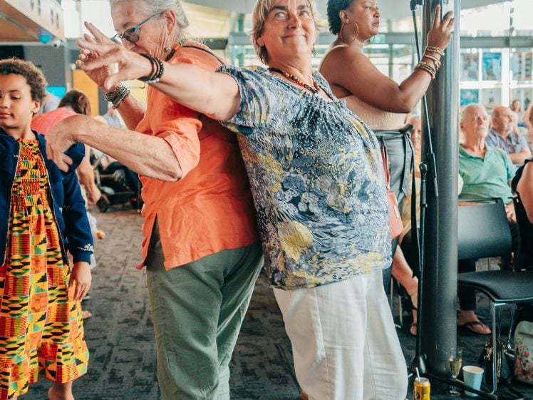 Image of Two older women stand back-to-back, smiling and posing playfully at an indoor event. Other people are seated or standing around them, creating a lively, cheerful atmosphere.
