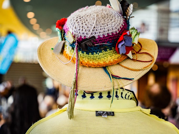 Image of A person wearing a colourful crocheted hat decorated with flowers, feathers, and accessories, viewed from behind. The person also wears a light yellow jumper with a patterned collar. The background is blurred.