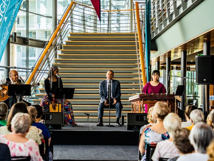Image of A musical group performs on a stage in front of a seated audience, with a wooden staircase and large windows in the background. The group includes a guitarist, two women musicians, and a man in a suit sitting centre stage.