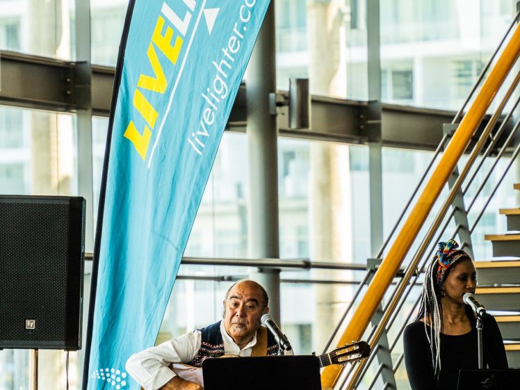 Image of A man plays guitar and sings beside a woman on percussion at an indoor event, with a blue LiveLighter banner and audience members visible in the foreground.