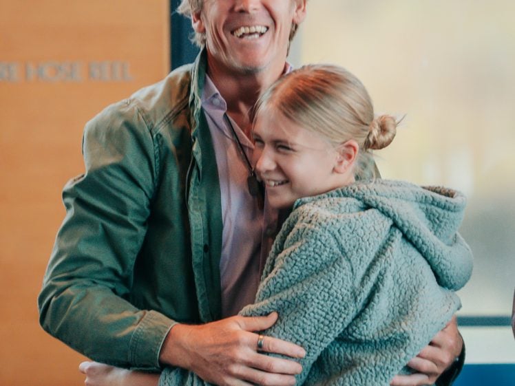 Image of A man and a young woman smile and embrace each other indoors. They both wear green jackets, and the background includes a door and a partially visible sign.