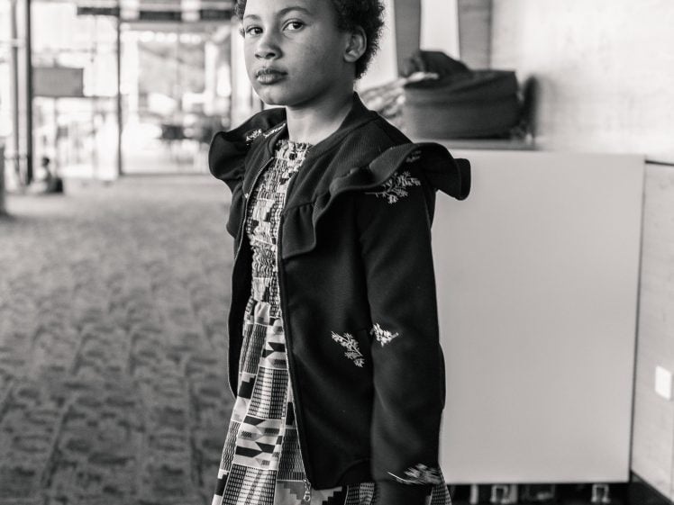 Image of A young girl with short curly hair walks in a corridor, wearing a patterned dress and a jacket. She looks confidently at the camera. The image is in black and white.