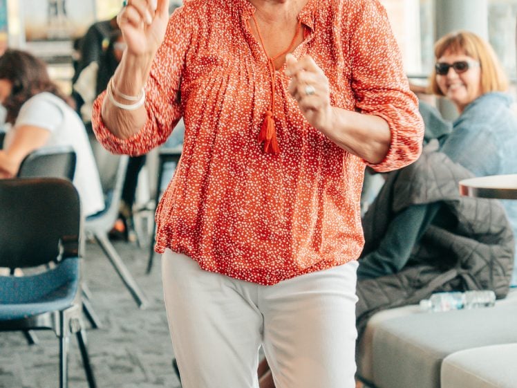 Image of An older woman with grey hair and glasses, wearing a red blouse and white trousers, smiles and gestures as she walks indoors in a modern, sunlit lounge area with other seated people in the background.