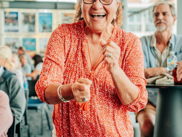 Image of An older woman wearing glasses, a red patterned blouse, and white trousers smiles excitedly and gestures with her hands in a lively, crowded indoor setting. Other people are visible in the background.
