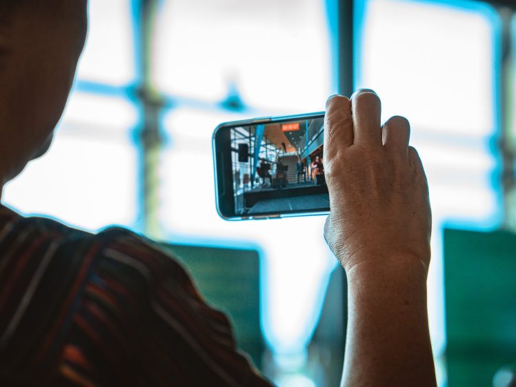 Image of A person holds up a mobile phone, taking a photo or video of a modern, glass-walled indoor setting, with blurred blue and green background elements visible through large windows.