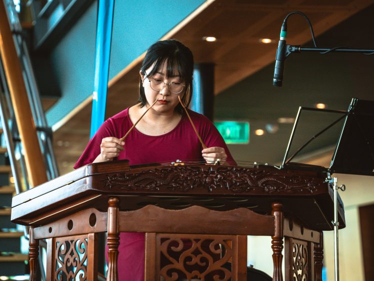 Image of A woman wearing glasses and a red dress plays a traditional stringed instrument with mallets. She stands behind a carved wooden table, with a microphone and sheet music nearby, in a modern indoor setting.