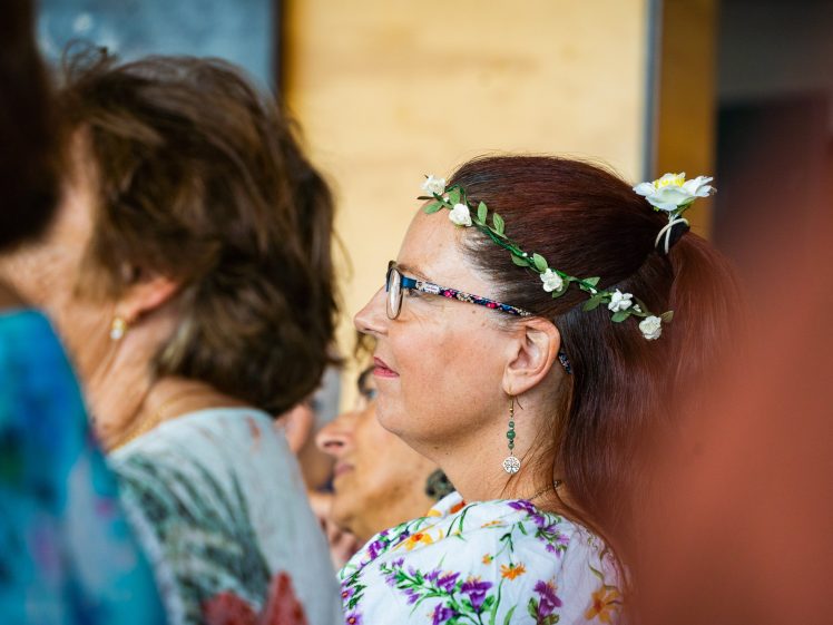 Image of A woman with glasses and a floral headband sits among a group of people. She wears floral-patterned clothing and earrings, and her brown hair is tied back. The atmosphere appears to be calm and attentive.
