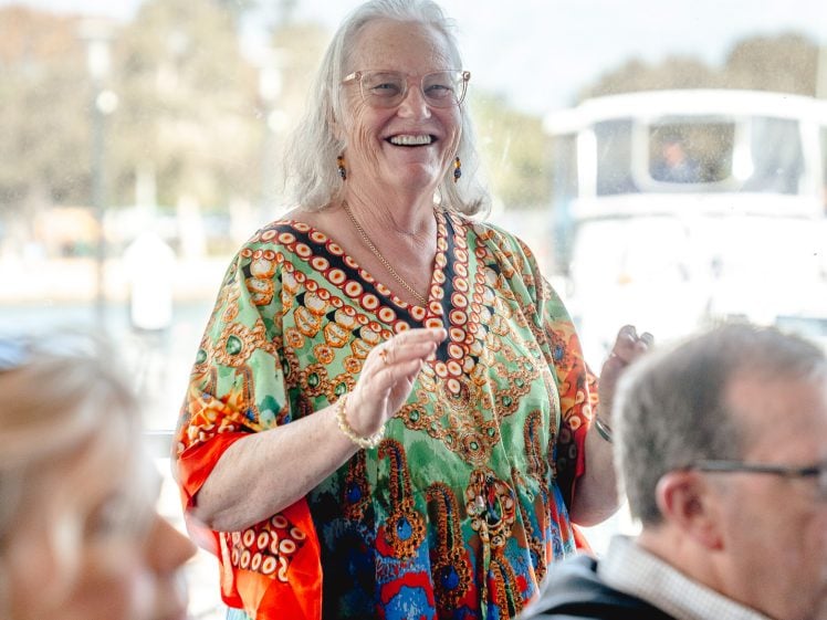 Image of An older woman with long white hair and glasses smiles and gestures while standing. She wears a colourful, patterned dress. People are seated nearby, and a boat is visible outside the window behind her.
