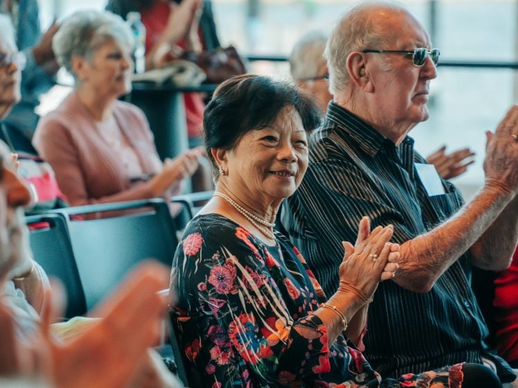 Image of A group of older adults sits in rows of chairs, clapping and smiling. The woman in the foreground wears a floral top and pearl necklaces, while the man next to her wears sunglasses and a striped shirt. The mood appears cheerful and engaged.