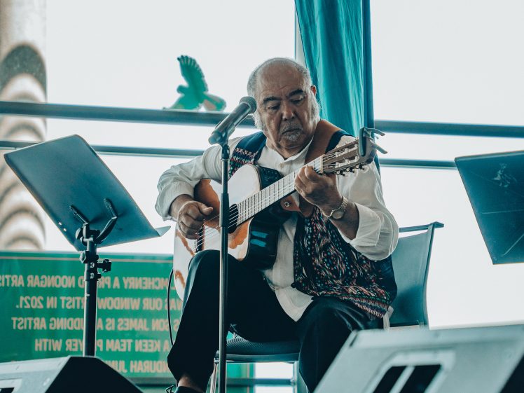 Image of An older man sits on stage playing an acoustic guitar and singing into a microphone. He wears a patterned waistcoat and white shirt. Music stands and a green sign are visible in the background.