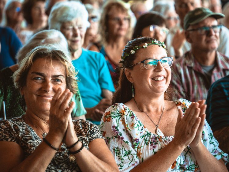 Image of Two women sit in a crowd, smiling and applauding. One wears a floral dress and a flower crown; the other has short hair and a patterned blouse. The background is filled with more people, all appearing to enjoy the event.