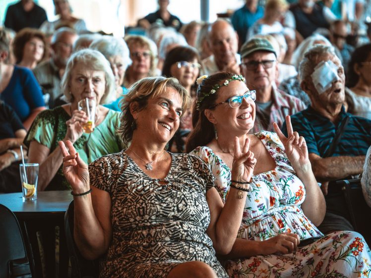 Image of Two women sitting in a crowd smile and make peace signs with their hands. The audience behind them consists mostly of older adults, some smiling, with one person drinking from a straw and another with an eye patch.