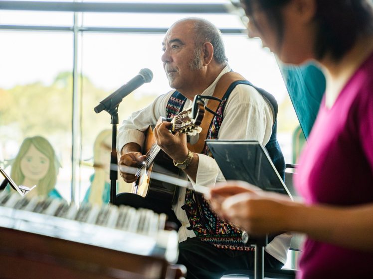 Image of A man sings into a microphone while playing guitar, seated indoors near large windows. A woman in the foreground holds papers, slightly out of focus. Colourful cartoon figures are visible in the background.