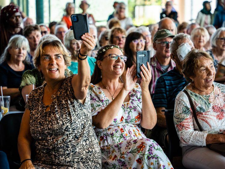 Image of A group of older adults sit closely together at an indoor event, smiling and laughing. Two women in the front row hold up mobile phones, appearing to record or take photos. The atmosphere is lively and cheerful.