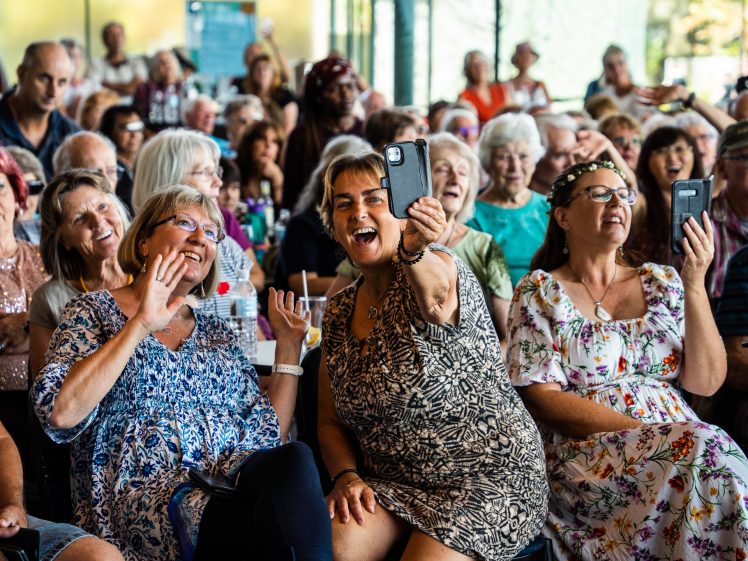 Image of A large group of older adults sit closely together at an indoor event. Several people smile, wave, and hold up mobiles to take photos or videos, creating a joyful and lively atmosphere.