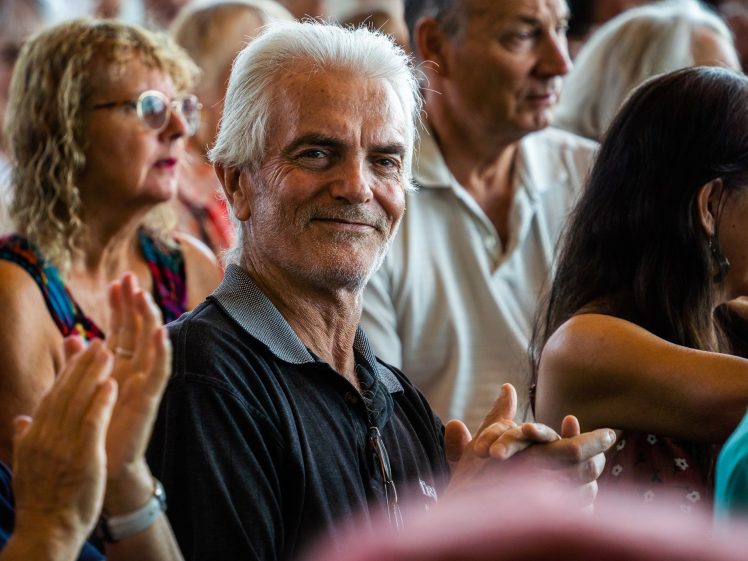 Image of An older man with white hair and a black polo shirt smiles whilst sitting among a crowd of people at an indoor event. Others around him are clapping and appear to be paying attention to something on stage.