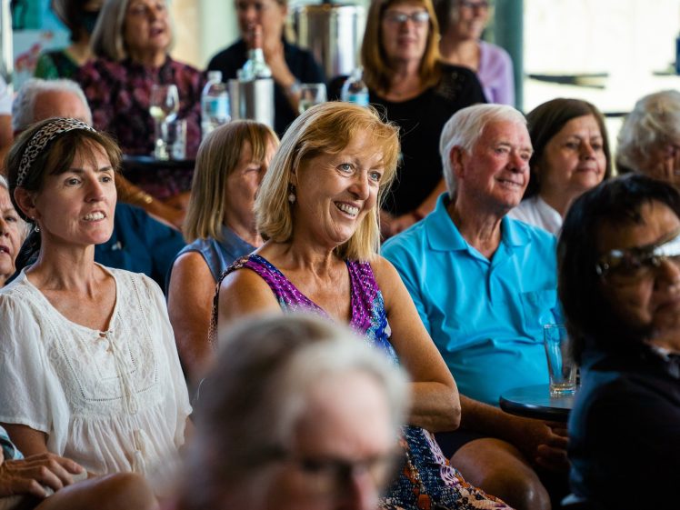 Image of A group of older adults sit closely together indoors, smiling and listening attentively. Most are women, and the atmosphere appears warm and engaging, suggesting they are enjoying a presentation or event.