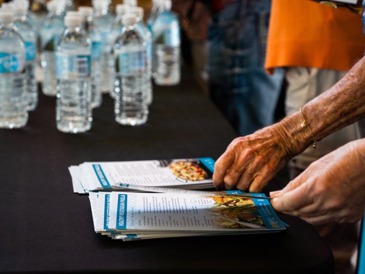 Image of A close-up of an elderly person's hands picking up leaflets from a table covered with bottled water and informational pamphlets at an indoor event.