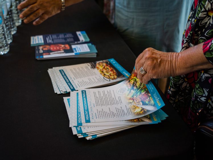 Image of A person picks up a stack of healthy eating leaflets from a table, where more leaflets, bottled water, and another person's hands are visible. The scene suggests an informational or community event.