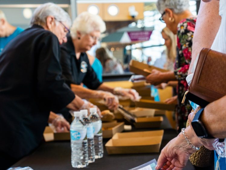 Image of A group of older adults at a table, some handing out food in brown boxes, while a woman in the foreground looks at leaflets and flyers next to bottled water.