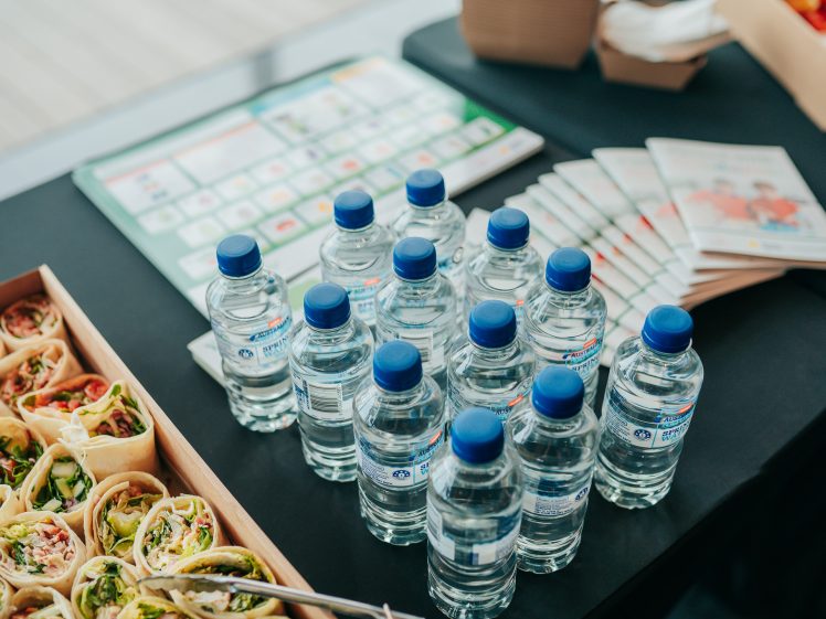 Image of Several bottles of water with blue caps are arranged on a black table next to platters of assorted wraps and some printed brochures or booklets in the background.
