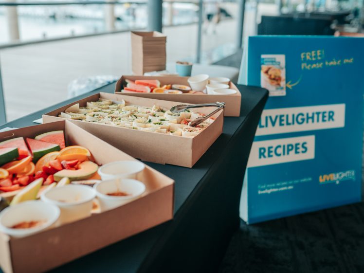 Image of A table with boxes of assorted sandwiches and fresh fruit slices next to small bowls of dip, positioned beside a blue sign that reads LiveLighter Recipes and FREE! Please take one.