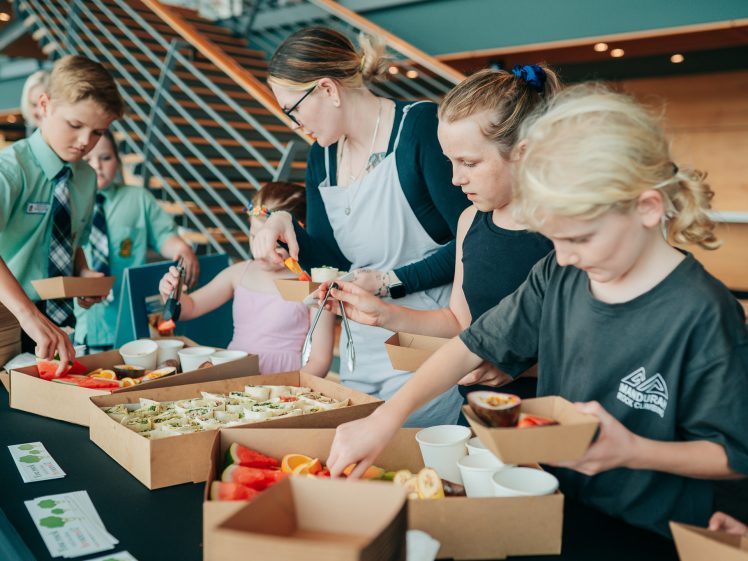 Image of A group of children and two adults select food from an assortment of fresh fruit and wraps, placing items into brown paper lunch boxes at a modern indoor venue.