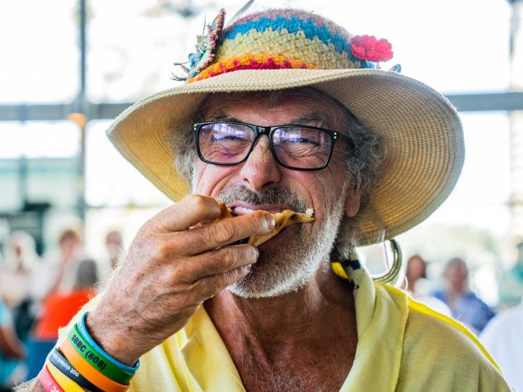 Image of An older man wearing a colourful straw hat, glasses, and rainbow wristbands smiles whilst eating pizza. He is dressed in a yellow shirt and appears to be enjoying himself at a lively indoor event.