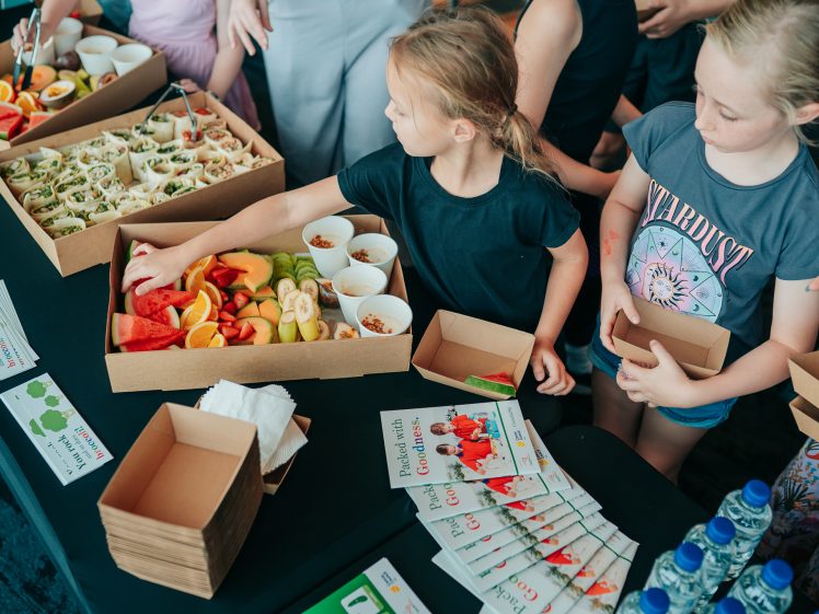 Image of Two young girls stand at a table with trays of wraps, fresh fruit, granola, and leaflets. One girl reaches for a piece of watermelon while holding an empty box. Bottles of water and serviettes are also on the table.