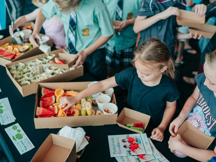 Image of Children gather around a table with lunchboxes filled with colourful fruits, vegetables, and wraps. Some children reach for food while others hold their boxes. Leaflets about healthy eating are displayed on the table.