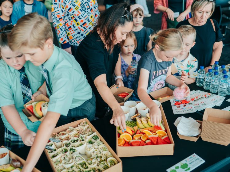 Image of A group of children and adults gather around a table filled with assorted healthy snacks, including wraps, fruit slices, and water bottles, as they select food items together.